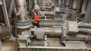 Man in an orange vest and white hard had stands in the middle of several large metal pipes