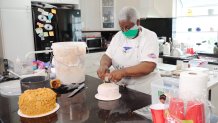 A Black woman with white hair stands in a kitchen, decorating a cake while wearing a white apron and a green mask over the lower half of her face