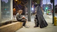 A man dressed as Batman engages in conversation with an older man seated on a low concrete wall next to a downtown building at night.
