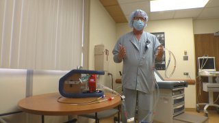 Man in light blue scrubs stands next to a table with a metal box and tubes.