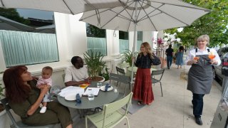 In this Saturday, June 19, 2021, photo, Caroline Styne, owner and wine director at The Lucques Group, standing under umbrella, welcomes back regular customers, Chris Anokute with his wife Jasmine and their 9-month-old son, Phoenix, at the A.O.C.