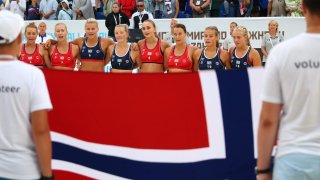 Members of Norway's Women's Beach Handball team stand in a line behind their flag.