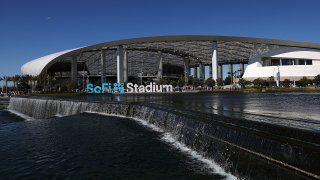 An exterior view of Los Angeles' SoFi Stadium, taken in daylight with a water feature and pond in front of the stadium.