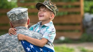 Military father hugs son holding American flag.