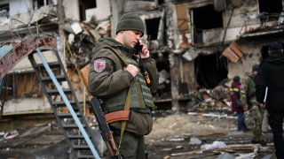A Ukrainian serviceman talks on a smartphone in front of a damaged residential building at Koshytsa Street, a suburb of the Ukrainian capital Kyiv, where a military shell allegedly hit, on February 25, 2022.