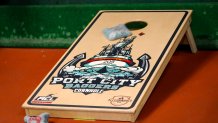 a bean bag slides up a cornhole board with the "port city baggers" logo that features a ship, an anchor, and a pirate throwing a bean bag.