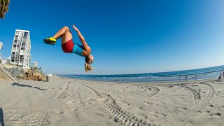 A woman doing a flip into the air off of a cement wall onto the sand of the beach while working out in the spring in the city of San Diego, California