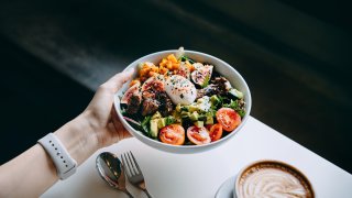 Close up of woman’s hand holding a bowl of fresh beef cobb salad, serving on the dining table. Ready to enjoy her healthy and nutritious lunch with coffee. Maintaining a healthy and well-balanced diet. Healthy eating lifestyle