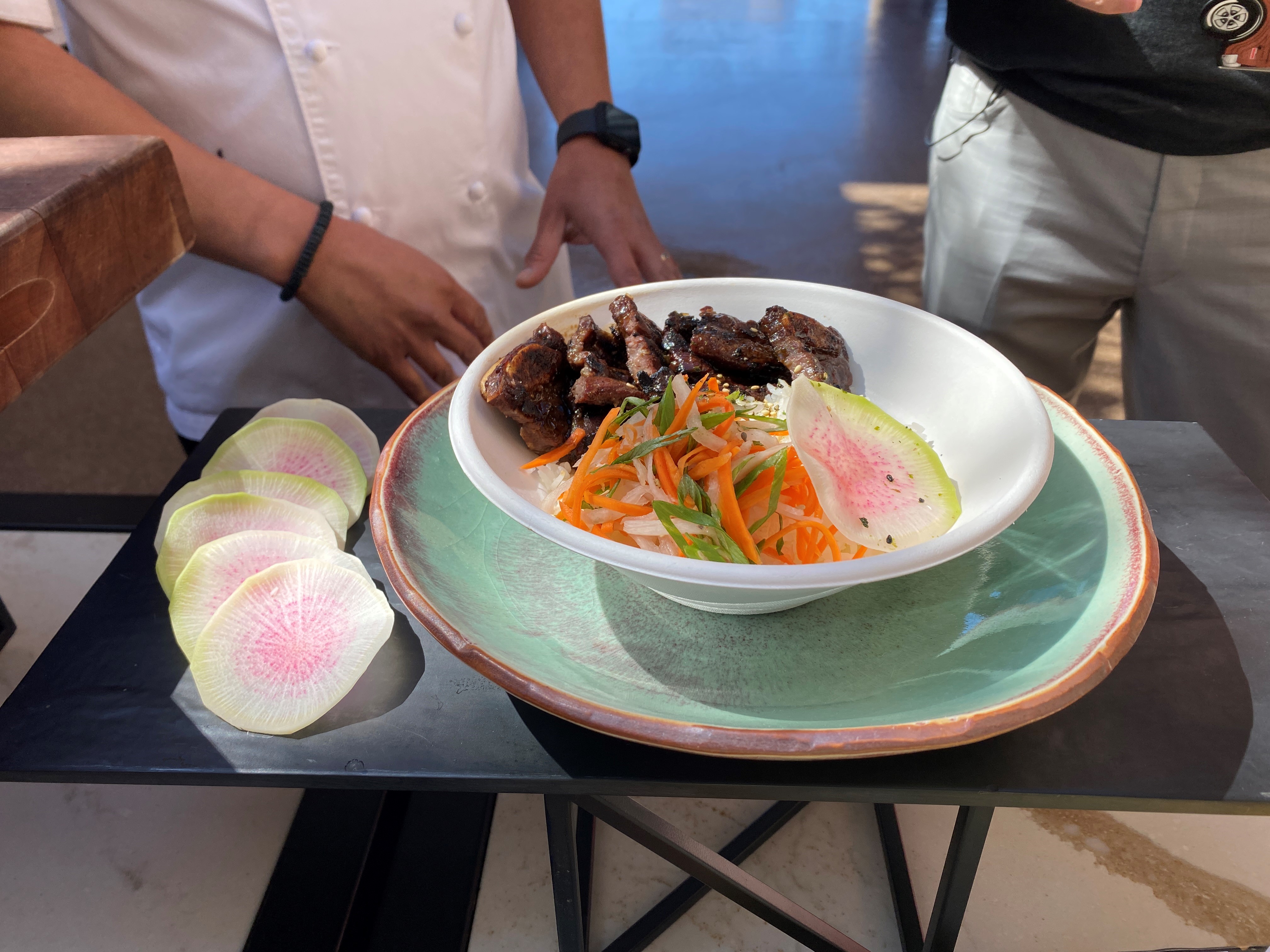 The Hawaiian Short Rib Bowl is a marinated beef short rib served with steamed rice, green onions, pickled daikon and carrot, furikake, and big wave sauce in the Kona Big Wave Patio in Section 227.
