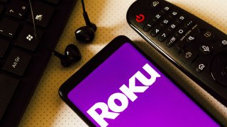 The logo of Roku is seen displayed on a smartphone next to a TV remote.