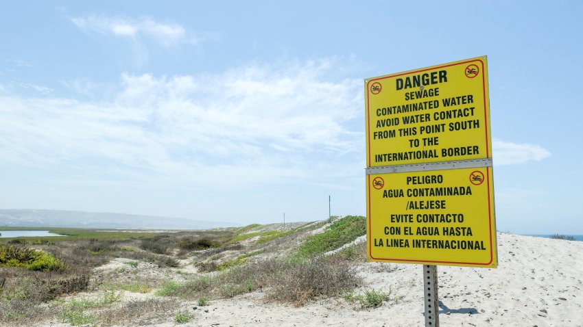 Bilingual warning sign near the mouth of the Tijuana River and Pacific Ocean. Text: DANGER, Sewage contaminated water. Avoid water contact from this point south to the international border. Identifiers removed.