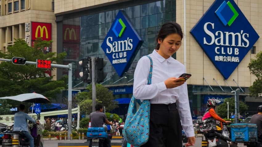 SHENZHEN, CHINA – APRIL 12: A woman checks her smartphone while walking past a busy intersection in front of a Sam’s Club membership store and a McDonald’s restaurant on April 12, 2025 in Shenzhen, China.