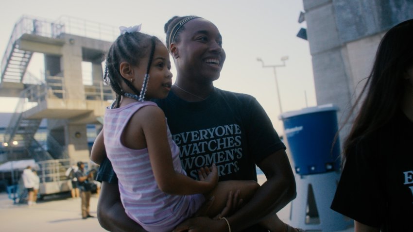 Olympian Simone Manuel wears a shirt that says "Everyone watches women's sports" and holds a young Black girl in her arms as they enter a pool area