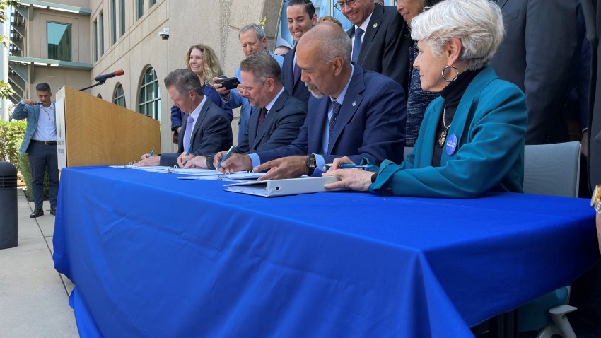 4 people sign papers while more people watch from behind.